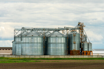 Fototapeta na wymiar next to the plowed agricultural field installed silver silos on agro manufacturing plant for processing drying cleaning and storage of agricultural products, flour, cereals and grain. Granary elevator