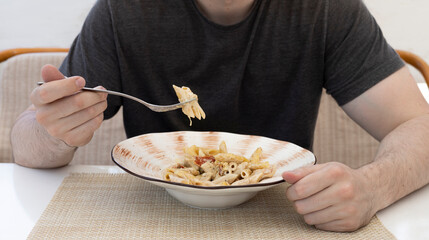 A young man eats Italian pasta from a beautiful plate at a white served table in a cafe.