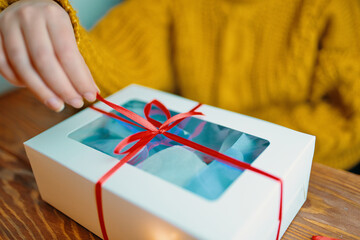 Woman in knitted sweater opens festive gift. Box with red ribbon on wooden table. Christmas or Birthday.