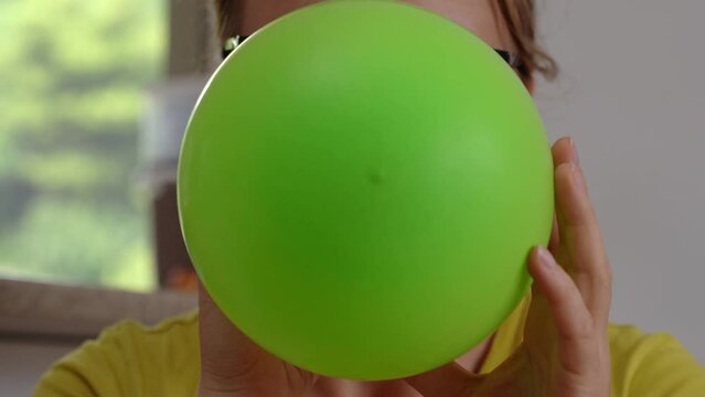 A close-up of a girl in glasses and a yellow T-shirt inflates a green ball and covers the entire frame with it.