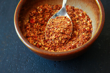 close-up of red dry chili powder in a brown ceramic bowl with a spoon on a dark background with space for text, red chili powder as cooking ingredient