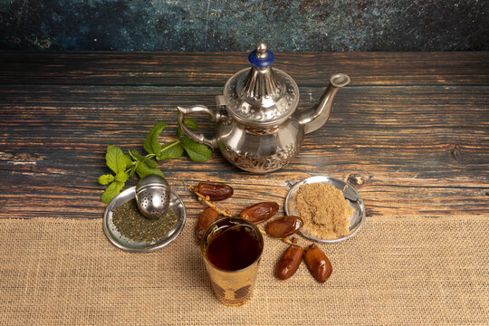 Arabic patterned cups and teapot filled with tea and mint or mint with dates, tea and cane sugar on two plates on a wooden table with a beige cloth and a stained cement-like background. 