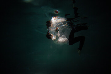 Fototapeta na wymiar Sink. A young guy in a white shirt falls into the water, a photo from under the water. The concept of falling down, diving to the depth, contrasting dark photo