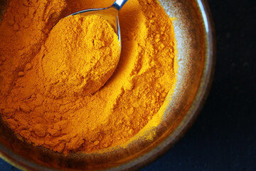 close-up of yellow turmeric spice powder with a spoon in a  brown ceramic bowl on dark background,...