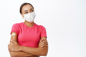 Smiling woman with patch on shoulder, did coronavirus vaccine, wears medical face mask, looks at banner, text or logo, stands over white background