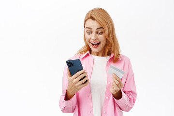 Mobile shopping, online application concept. Smiling young woman holding smartphone and credit card, looking excited, standing over white background