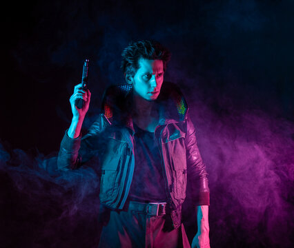 A guy in a cyberpunk image, holding a gun in his hands. Futuristic character in smoke. A young man in neon lighting on a black background