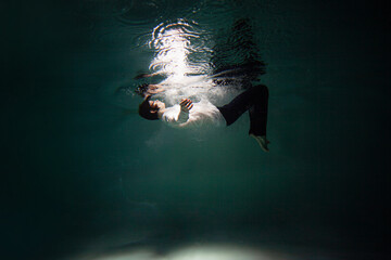 Sink. A young guy in a white shirt falls into the water, a photo from under the water. The concept of falling down, diving to the depth, contrasting dark photo - 514532880