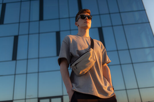 Portrait of handsome tourist hipster man in sunglasses with bag handy, standing guy near the building in downtown. Calm relaxing moment, summer vacation concept