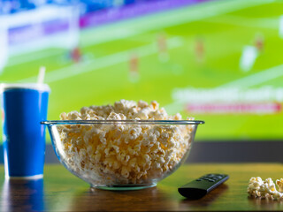 A TV remote control, a large bowl of popcorn and a soda drink in a plastic glass against the...