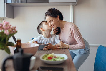 Portrait of cute small boy having breakfast and kissing his mom at table in kitchen.