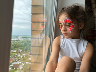 a little girl sits by the window and dreams and thinks. beautiful face art butterfly on a face of child