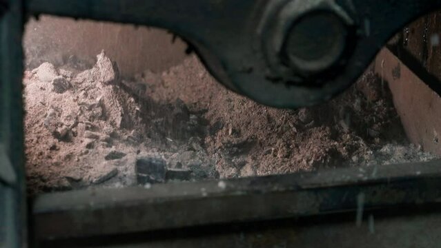 Ash falls from the firebox of a wood-fired boiler into an ash container from below, close-up
