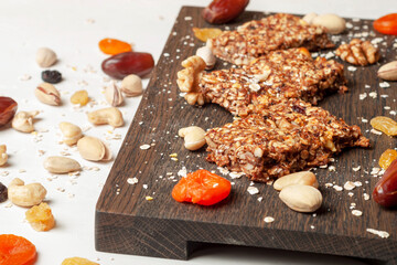 bar with flakes, nuts and dried fruits on a brown wooden board with ingredients scattered side by side on a gray background. the bars lie side by side with each other, the board is partially visible.