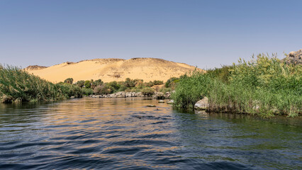 The riverbed bends between banks overgrown with green vegetation. An orange sand dune against a clear blue sky. Reflection on calm water. Egypt. Nile. Aswan
