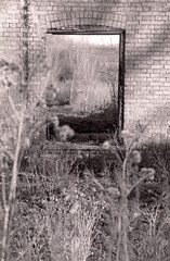 Derelict barn window no glass or frame. Old rough brick exterior. Dried grasses in foreground and background. Sunlight and shadow. Photo taken1998. Black and white film. Scanned negative. Sepia tone