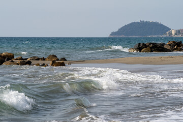 Beach on the Italian Riviera with sun, sea and stones in the sea water. Rough Sea and Foamy Waves on Shore at Mediterranean Coast in Sunrise