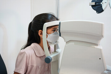 Ophthalmologist examining the eyes of an Asian girl in a clinic. They wear protective face masks.
