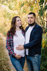 Fototapeta premium Pregnant woman, mom-to-be, stands next to dad in the autumn forest and hugs. Gently touch by hand of the stomach with the future child. Pregnancy, love. High quality shot.