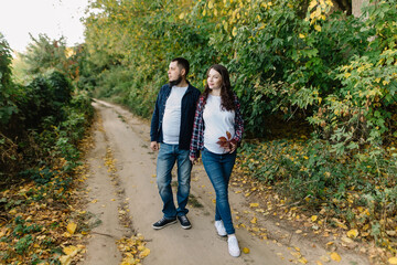 Obraz premium Beautiful young couple of future parents stand surrounded by autumn nature in the countryside outside the city and look into the distance. Pregnant woman holds an autumn leaf near her stomach. High