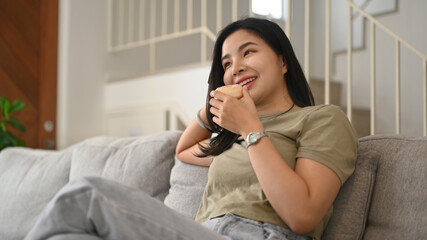 Pretty young woman eating donut and watching television on comfortable sofa at home