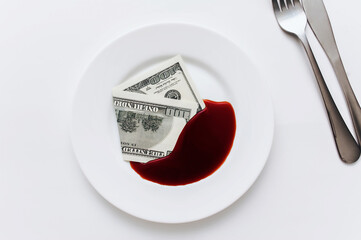 A hundred-dollar bill lies in a white plate near to blood-like soy sauce with the fork, knife on...