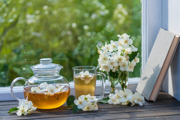Hot herbal tea in glass teapot, cup and beautiful bouquet of jasmine flowers on windowsill at...