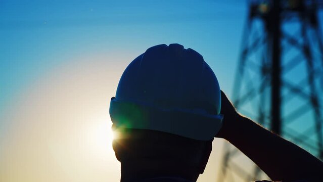 Silhouette male builder in helmet on field at tall telecommunications tower. Construction worker viewing construction site before starting construction work. Rear view. Removing head dress hand.