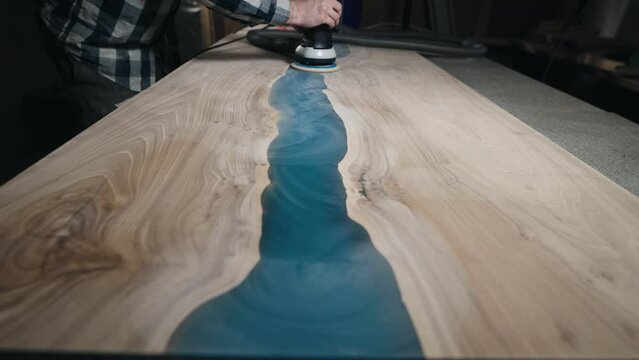 The carpenter processes the surface of a wooden tabletop with a grinder. Slab filled with epoxy resin.