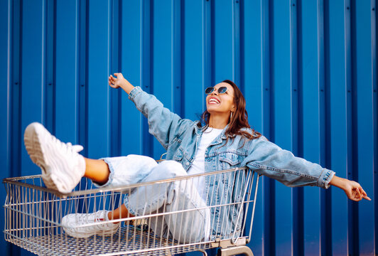Happy woman with shopping cart having fun near color wall.  Consumerism, sale, purchases, shopping, lifestyle concept.