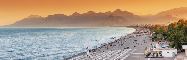 Naklejka premium Sunset panoramic view of scenic and popular Konyaalti beach in Antalya resort town. Majestic mountains with haze in the background. Vacation and holiday in Turkey