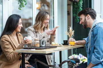 Group of cool friends sharing a brunch together while talking and looking the smartphone on the healthy coffee shop terrace.