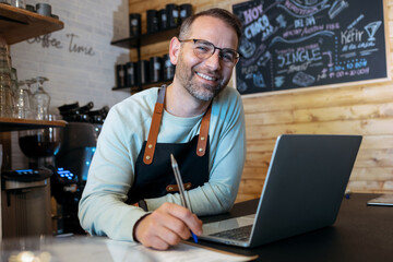 Handsome mature owner bakery working while analyzing report for order delivery with laptop in a...