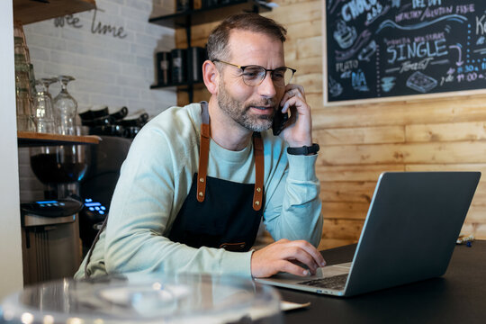 Handsome Mature Owner Bakery Working While Analyzing Report For Order Delivery With Laptop In A Pastry Shop.