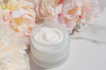 Cosmetic cream with fresh flowers of pink peony blossoms on light marble background. Dermatologic, components for cosmetics.