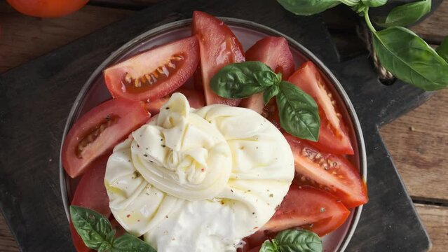 Salad with tomatoes and burrata cheese with basil and olive oil in a plate on wooden background. Top view. Italian food. 4k video
