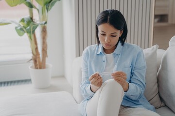 Unhealthy young woman suffer from high body temperature, holding thermometer sitting on sofa