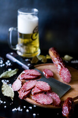 Salami and beer. Salami is cut into slices with a large knife. Smoked sausage. Salami sausage on a wooden board ona a black background. sausage with spices. A glass of beer with a sausage.