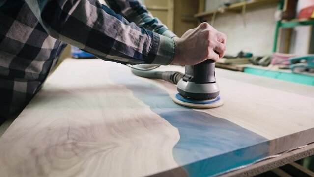 The carpenter processes the surface of a wooden tabletop with a grinder. Slab filled with epoxy resin.