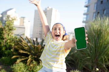 Joyful young girl holding smartphone in her hand. Green screen is turned towards viewer. Teenager with phone in park sitting on stylish bench. Selective focus. Emotions, connection, digital concept