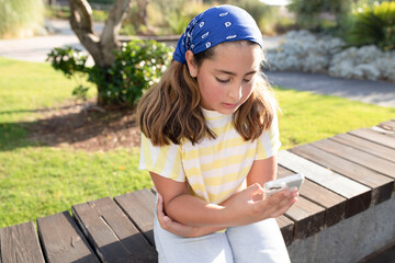 Teenager with smartphone outside. Evening sun light. Stylish girl in bandana is concentrating on texting, looking for content on Internet. Concept of blogging, follower, kids and gadgets, online, life