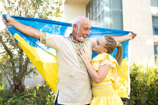 Elderly Man In Ukrainian Authentic Clothes With Little Girl In Yellow Dress Holding Flag Of Ukraine. Grandfather And Granddaughter. Outside. Concept Of Refugees, Support, Family, Together, War, Peace.