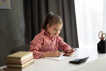 Caucasian child schoolgirl holding phone distance learning class using mobile application, watching online tutoring lesson, making notes studying at home in classroom.