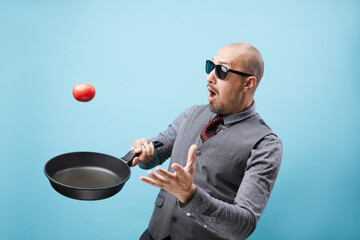A  funny chef man in suit holding frying pan and juggling tomato isolated over blue wall background.