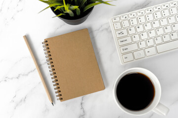 Office desk with brown notepad, cup of coffee, pencil and computer keyboard on top of marble desk
