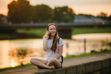 A girl talking on her cell phone while sitting on the riverbank during sunset.
