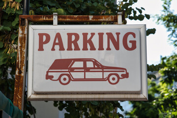 A retro-style sign saying 'parking' hanging on a rusty pole with the leaves of a tree in the...