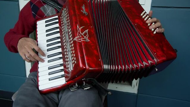 A well-dressed man is sitting on a chair and playing a red accordion traditional musical instrument on the terrace of the house