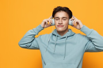a happy man in a blue hoodie stands on an orange background listening to music with headphones holding them with his hands closing his eyes with pleasure