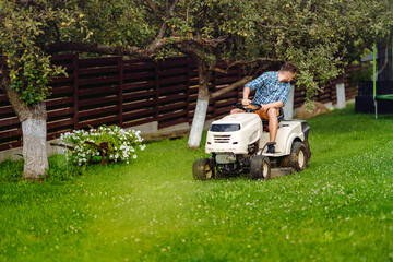 Professional gardener driving a riding lawn mower in a garden. Industrial landscaping works. - obrazy, fototapety, plakaty